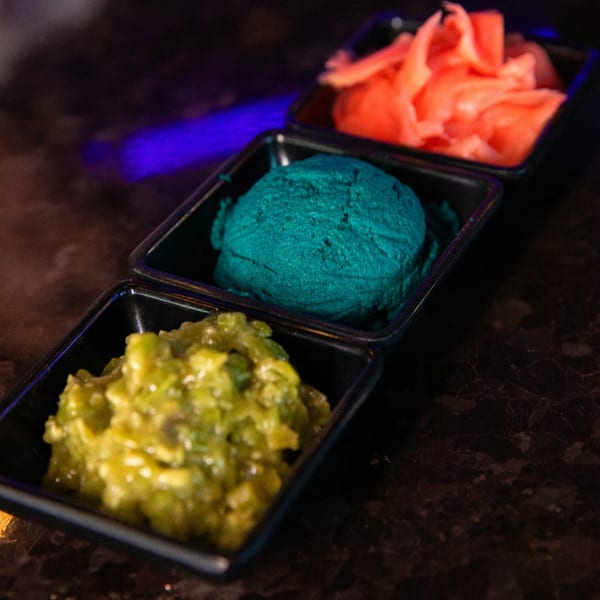 Three small black dishes containing pickled ginger, blue wasabi, and green wasabi on a dark surface.