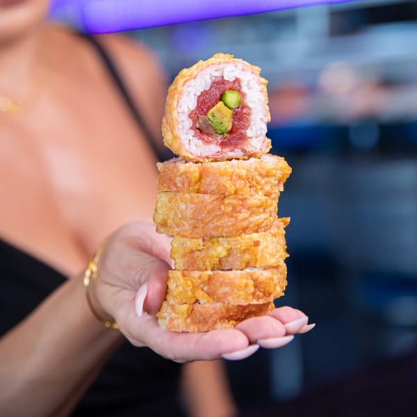 A hand holding a tall stack of crispy, deep-fried sushi rolls with tuna and avocado.
