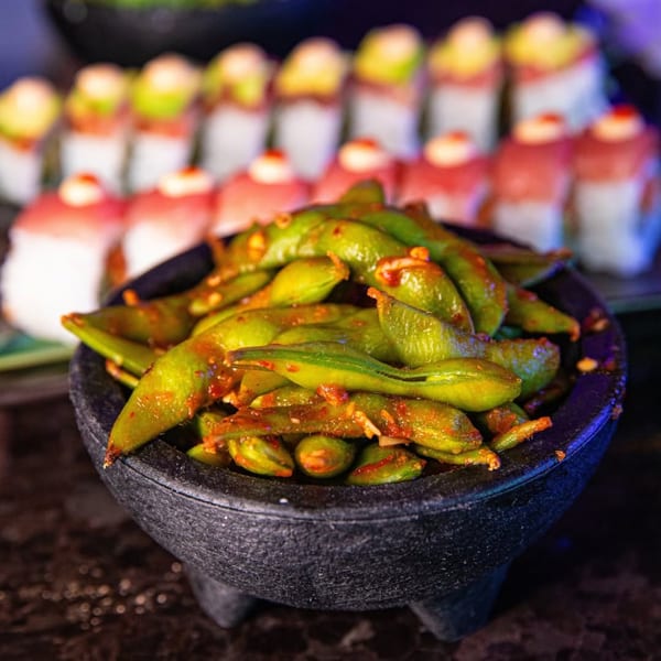 Spicy edamame in a dark stone bowl, with rows of sushi rolls blurred in the background.