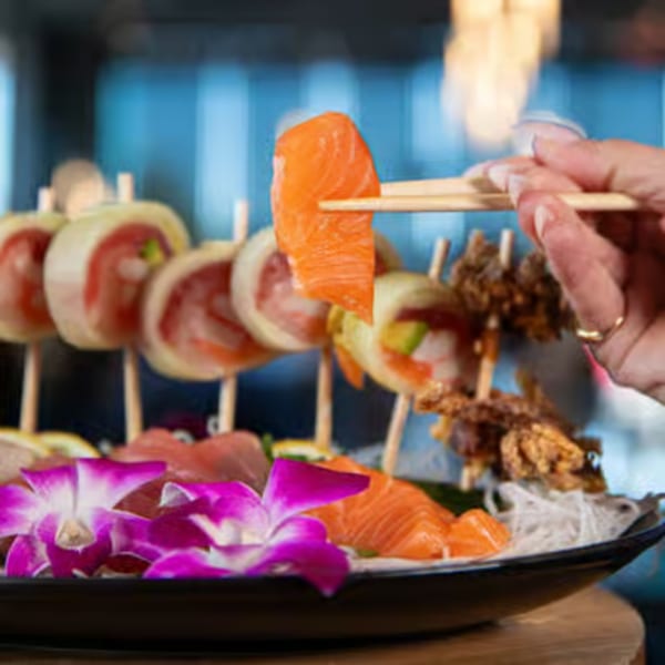 Chopsticks holding a piece of salmon sashimi over a platter of sushi and purple orchids.