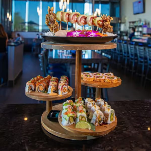 A three-tiered wooden stand holds various sushi rolls and skewers, with a restaurant interior in the background.